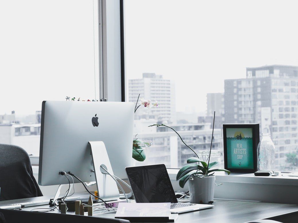 Two Apple computers on a desk inside an office next to a window overlooking a cityscape.